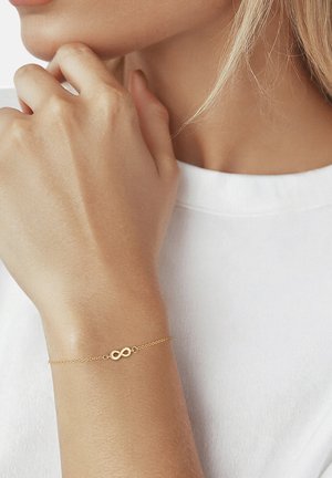 Delicaat gouden kettingarmband met een oneindigheidssymbool, gedragen om de pols tegen een witte overhemd en een lichte huidskleur.