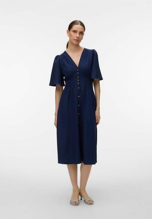 Nő sötétkék, gombos, térdig érő ruhában, rövid, felfújt ujjakkal és ezüst nyitott orrú magassarkúban, egyszínű háttér előtt állva.