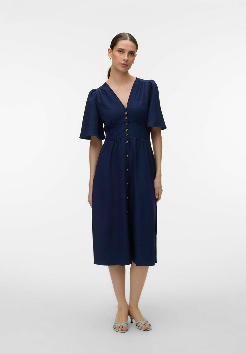 Femme portant une robe bleu marine boutonnée, longueur genou, avec des manches courtes bouffantes et des talons argentés à bout ouvert, debout devant un fond uni.