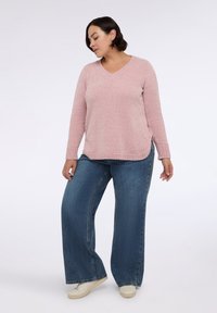 Helles rosa strukturiertes Sweatshirt mit V-Ausschnitt und seitlichen Schlitzen, kombiniert mit blauen, weit geschnittenen Jeans und beigen Turnschuhen. Lässige Outfit-Zusammenstellung.