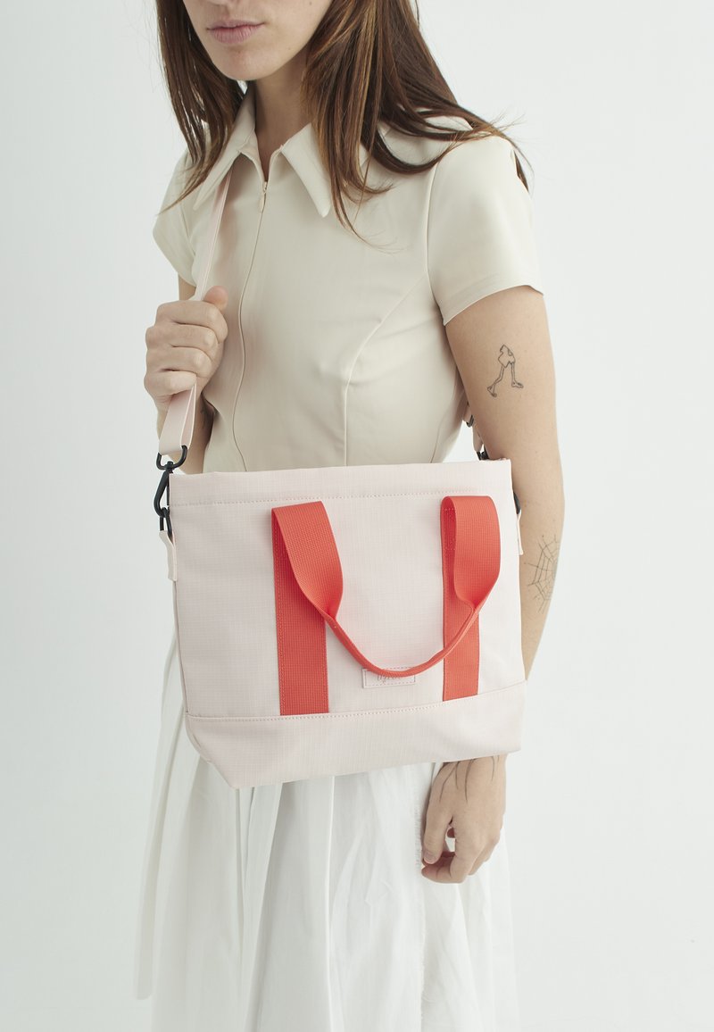 Rosa stof tote bag med røde håndtag, firkantet design, flad bund og en tekstureret overflade. Har en aftagelig skulderrem.