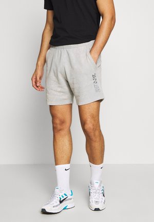 Homme portant un short gris clair avec le texte "Just Do It", des chaussettes Nike blanches, des baskets Nike blanches et bleues, et un t-shirt noir.