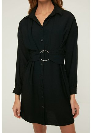 Vrouw draagt een zwarte jurk met knopen, lange mouwen en een brede riem met een grote zilveren ronde gesp in de taille.