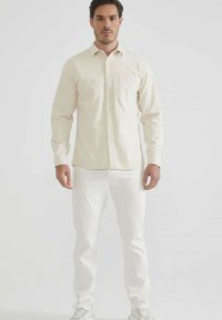 Homme faisant face, portant une chemise beige à boutons, un pantalon blanc et des baskets blanches, devant un arrière-plan léger et uni.