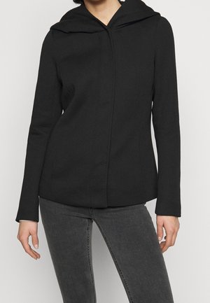 Osoba w dopasowanej czarnej bluzie z kapturem i długimi rękawami oraz ciemnoszarych dżinsach, stojąca na tle jednolitego białego tła.