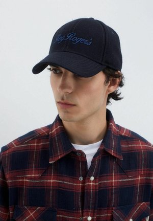 Cappello navy con ricamo "Rogers" sulla parte anteriore, abbinato a una camicia a quadri rossa e navy con bottoni. Texture del tessuto naturale visibile.
