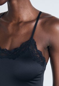 Gros plan sur le torse supérieur d'une personne portant un caraco noir avec des dentelles et de fines bretelles contre un fond uni.