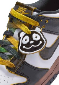 Nike-sneakers med en vit läderöverdel, svarta accenter, snören med gul och grön gradient och en pandaface-lace charm.