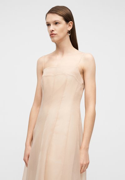 Γυναίκα με καστανά ίσια μαλλιά που φορά αμάνικο, ανοιχτό μπεζ φόρεμα και χρυσούς κρίκους στα αυτιά, κοιτάζοντας ελαφρώς στο πλάι.