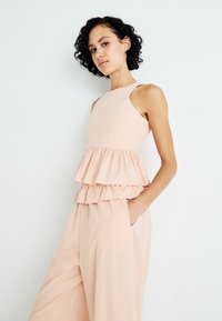 Vestido sin mangas de color durazno con un corsé ajustado y dos volantes en la cintura. Tela ligera, bolsillos laterales, corte relajado.