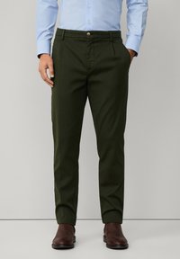 Pantaloni din bumbac verzi cu o croială elegantă, dotati cu buzunare frontale, fermoar și închidere cu buton. Purtați cu o cămașă bleumarin deschis și cizme maro.