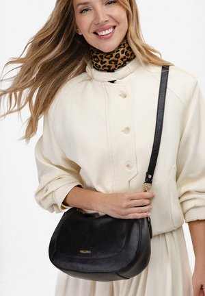 Vrouw die lacht, gekleed in een crème geknoopte jas en een leopardprint top, en een zwarte leren crossbodytas met gouden accenten vasthoudt.