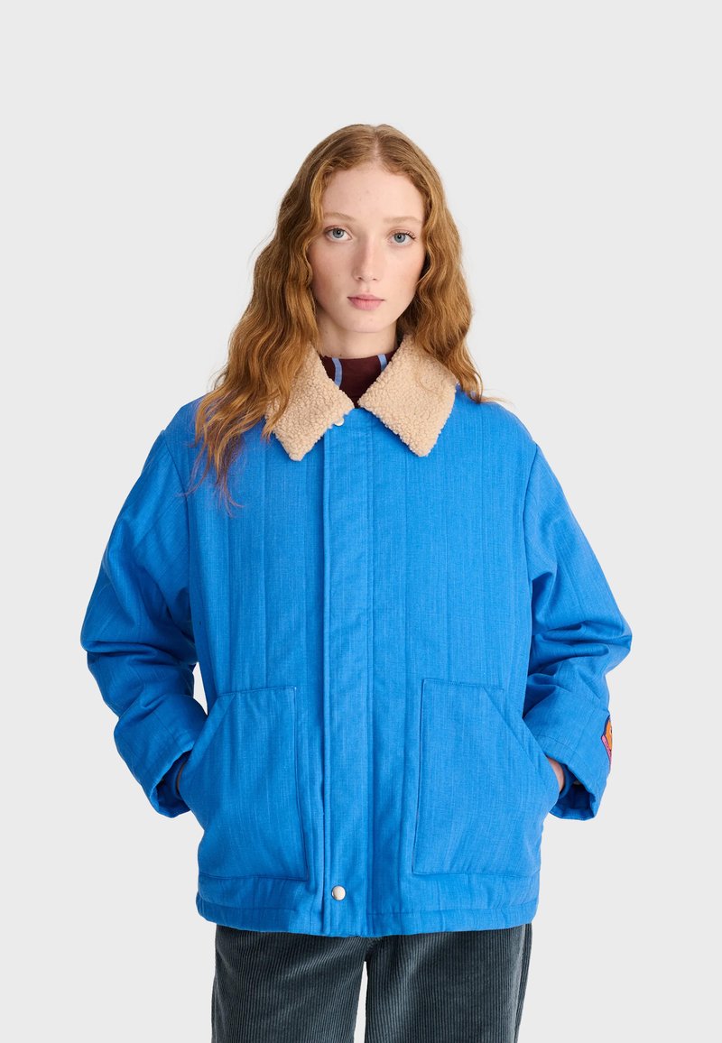 Jeune femme aux longs cheveux roux porte une veste bleue avec col sherpa beige et les mains dans les grandes poches avant, debout devant un fond uni.