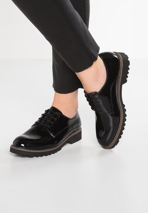 Chaussures à lacets noires brillantes avec semelles épaisses et crantées portées avec un pantalon noir retroussé sur un fond blanc uni.