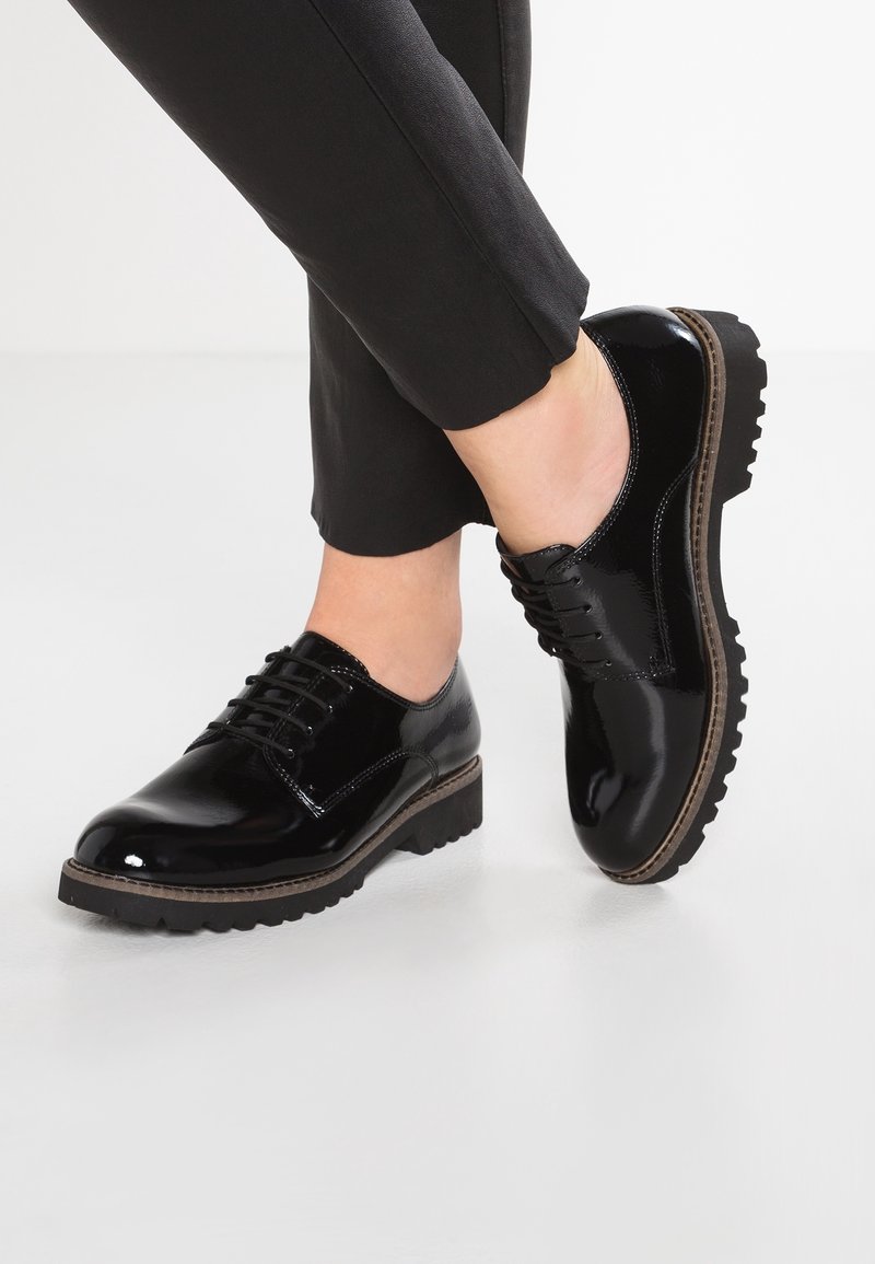 Chaussures à lacets noires brillantes avec semelles épaisses et crantées portées avec un pantalon noir retroussé sur un fond blanc uni.