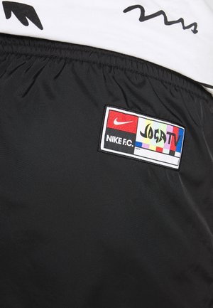 Czarna tkanina z naszytym prostokątnym łatką przedstawiającą czerwone logo Nike, tekst „"NIKE FC."” oraz kolorowe paski z tekstem „"JOGA TV"”.