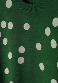 Pull vert en tissu texturé avec des pois blancs de tailles variées. Le col côtelé ajoute du détail au design.
