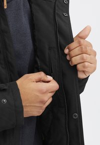 Svart jakke med glidelås-lomme på innsiden og trykknapplukking. Har en jevn tekstur og en tettsittende design, delvis åpnet.