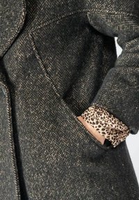 Mano parzialmente in tasca di un cappotto in lana grigio scuro a spina di pesce, con il polsino in tessuto leopardato che spunta dalla manica.