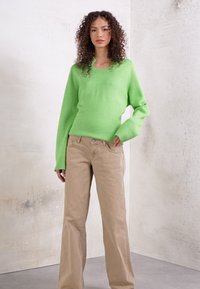 Suéter verde de malha suave com decote redondo e mangas longas, combinado com calças bege claras, ligeiramente largas, sobre um fundo neutro.