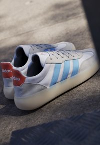 Par hvide sneakers med lyseblå striber og orange hælflapper, der hviler på en tekstureret grå overflade i sollys.