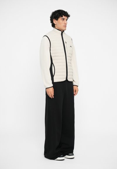 Padded vest i lys beige med høy krage og svarte sideakcenter. Bært over løstsittende svarte bukser, vist mot en hvit bakgrunn.
