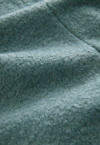 Měkká, strukturovaná látka v tlumené tyrkysové barvě s fuzzy povrchem a mírně vystouplým vzorem, vhodná na teplé oblečení nebo doplňky.