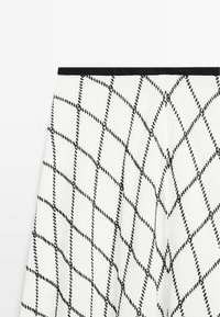 Hvid nederdel med et diamantmønster dannet af sorte syninger, med en sort talje. Stoffet ser tekstureret og struktureret ud.