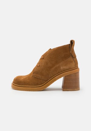 Botas de ante marrón hasta el tobillo con tacón bloque, que presentan una textura suave, punta redonda, altura hasta el tobillo y costuras decorativas en los laterales.