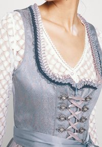 Dirndl traditionnel avec un corsage à motif bleu clair, décolleté froncé en marine, ferrures en argent et un détail de lacet en satin. Manches en dentelle blanche.