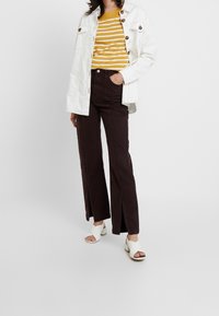 Giacca di jeans bianca con bottoni marroni, top a righe giallo senape, pantaloni larghi marroni scuri con spacchi frontali e sandali bianchi a slip-on.