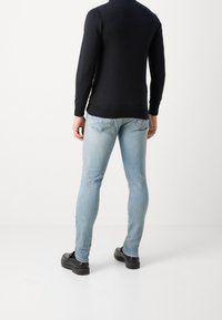 Schwarzes Langarmoberteil mit tailliertem Design, kombiniert mit hellblauen Slim-Fit-Jeans und schwarzen Schuhen, von hinten betrachtet.