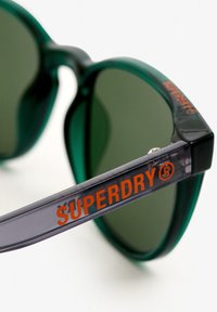 Zielone okulary przeciwsłoneczne o okrągłej oprawie, z przezroczystą szarością na ramieniu. Na ramieniu znajduje się napis "SUPERDRY®" w jaskrawej pomarańczowej czcionce.