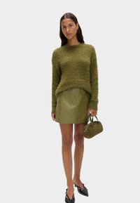Maglione verde oliva morbido con scollatura tonda, abbinato a una gonne in pelle verde oliva aderente. Tiene una piccola borsa verde. Scarpe nere a punta.