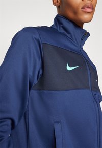 Mörkblå träningsjacka med hög krage, texturerad tyg och ljusblå Nike-logotyp på bröstet. Har dragkedja och sidofickor.