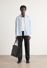 Lyseblå stripete skjorte med knapper, hvit tanktop, svarte bukser og en svart tote bag med preget logo; stilige sko fullfører antrekket.