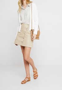 Gonna di jeans beige con bottoni davanti, abbinata a una camicia a quadri grigia chiara e a un cardigan bianco. Sandali marroni con cinturini e una borsa intrecciata completano l'outfit.