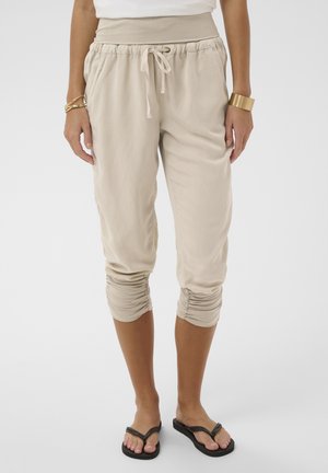 Vrouw draagt beige cropped broek met trekkoord en geplooide manchetten, wit topje, gouden armbanden en zwarte slippers, staand op witte achtergrond.