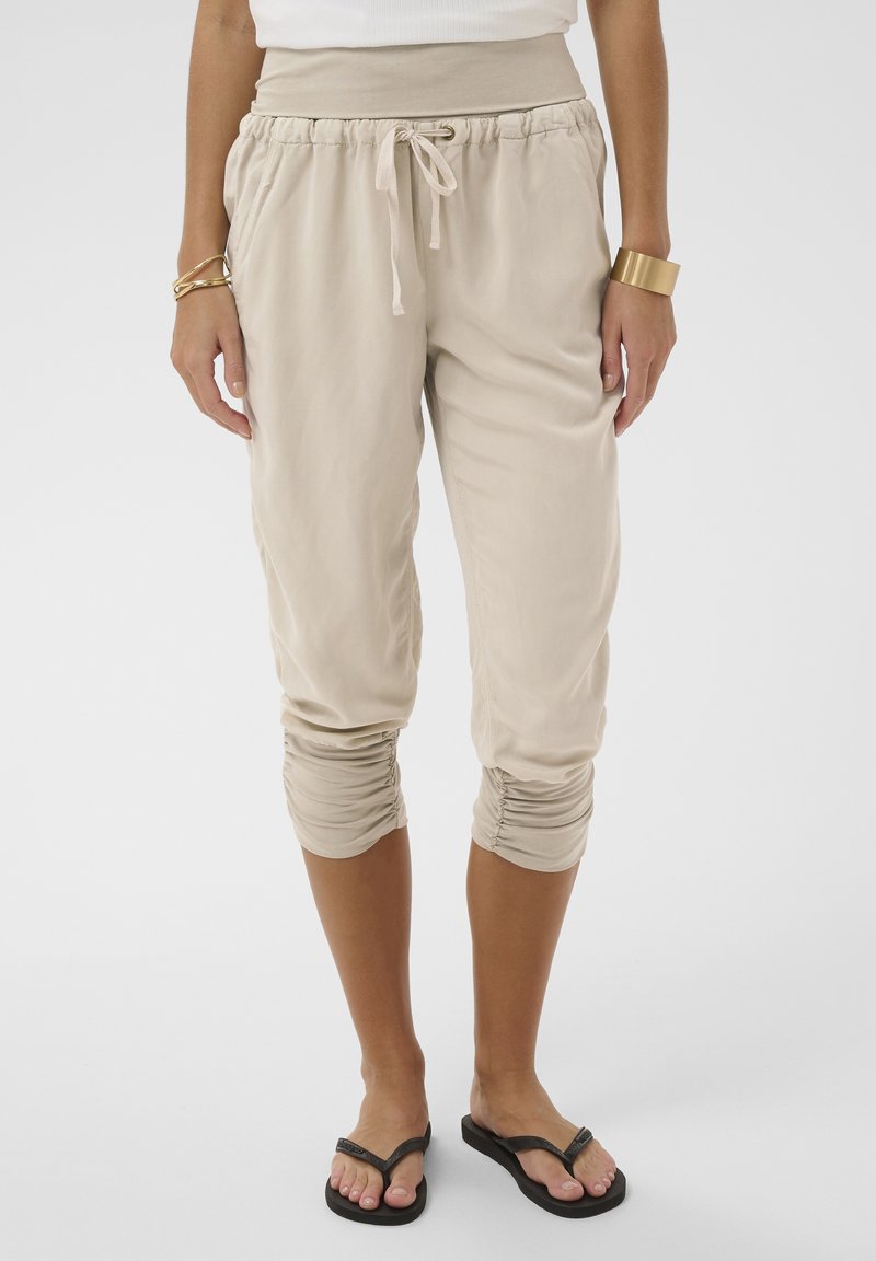 Donna che indossa pantaloni beige tagliati con coulisse e polsini arricciati, top bianco, braccialetti dorati e infradito nere, in piedi su sfondo bianco.