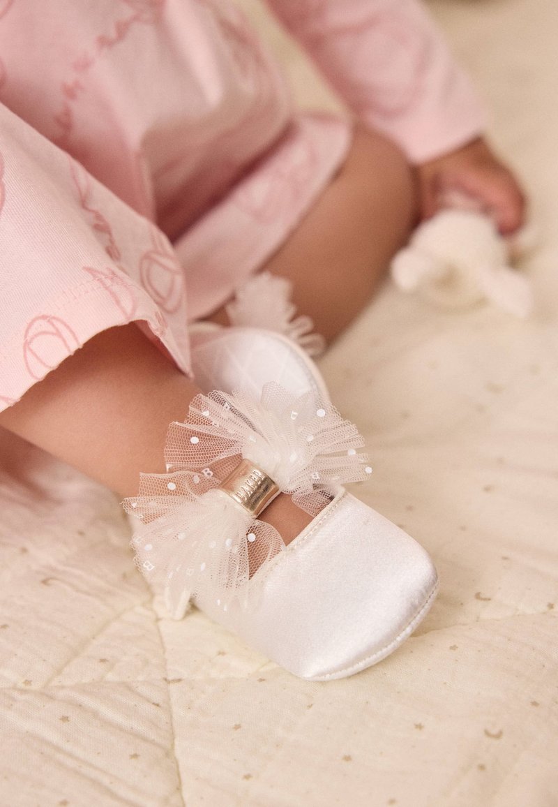 Scarpine da bambino in raso bianco con punta arrotondata, caratterizzate da un fiocco in tulle trasparente e strutturato decorato con piccoli pois bianchi e una fascia argentata.