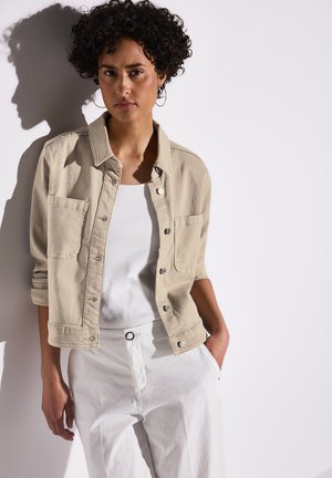 Veste en denim beige avec fermeture à boutons et deux poches avant. Portée par-dessus un débardeur blanc, assortie à un pantalon blanc, offrant une coupe décontractée.