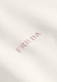 Biała tkanina o teksturowanej powierzchni z wypukłymi, pastelowymi różowymi literami tworzącymi napis "FRIEDA". Prosty, czysty design, bez dodatkowych wzorów.