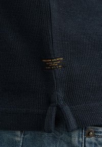 Marineblauer Strickpullover aus strukturiertem Material, mit seitlichen Schlitzen und einem Etikett, das "FREEDOM UNLIMITED" mit Spezifikationen trägt.