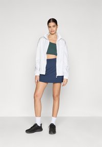 Hvid zip-up jakke, mørkegrøn crop top, blå mini nederdel og sorte sneakers. Funktioner inkluderer elastiske manchetter og et minimalistisk design.