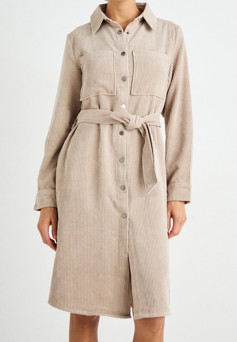 Robe chemise en velours côtelé beige avec manches longues, devant boutonné, deux poches poitrine, et ceinture assortie en tissu. Rayures verticales texturées.