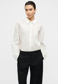 Chemise blanche à boutons avec col, manches longues et poche poitrine, associée à un pantalon noir ajusté. Texture lisse, design décontracté.