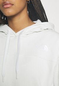 Sudadera gris claro con cordón, que presenta un pequeño logo bordado en blanco en el pecho. Tela suave con una textura lisa.