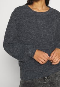 Mörkgrå stickad tröja med rund halsringning och långa ärmar. Har en texturerad tygkvalitet och ribbade muddarna, bärs över mörka jeans.