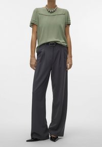 Blusa verde oliva a maniche corte con dettagli sottili, abbinata a pantaloni grigi a gamba larga, completata da una collana argento spessa e tacchi neri.
