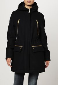 Zwarte gewatteerde jas met capuchon, gemaakt van zachte stof, met gouden ritsdetails op de borst en zakken, en een recht gesneden ontwerp.